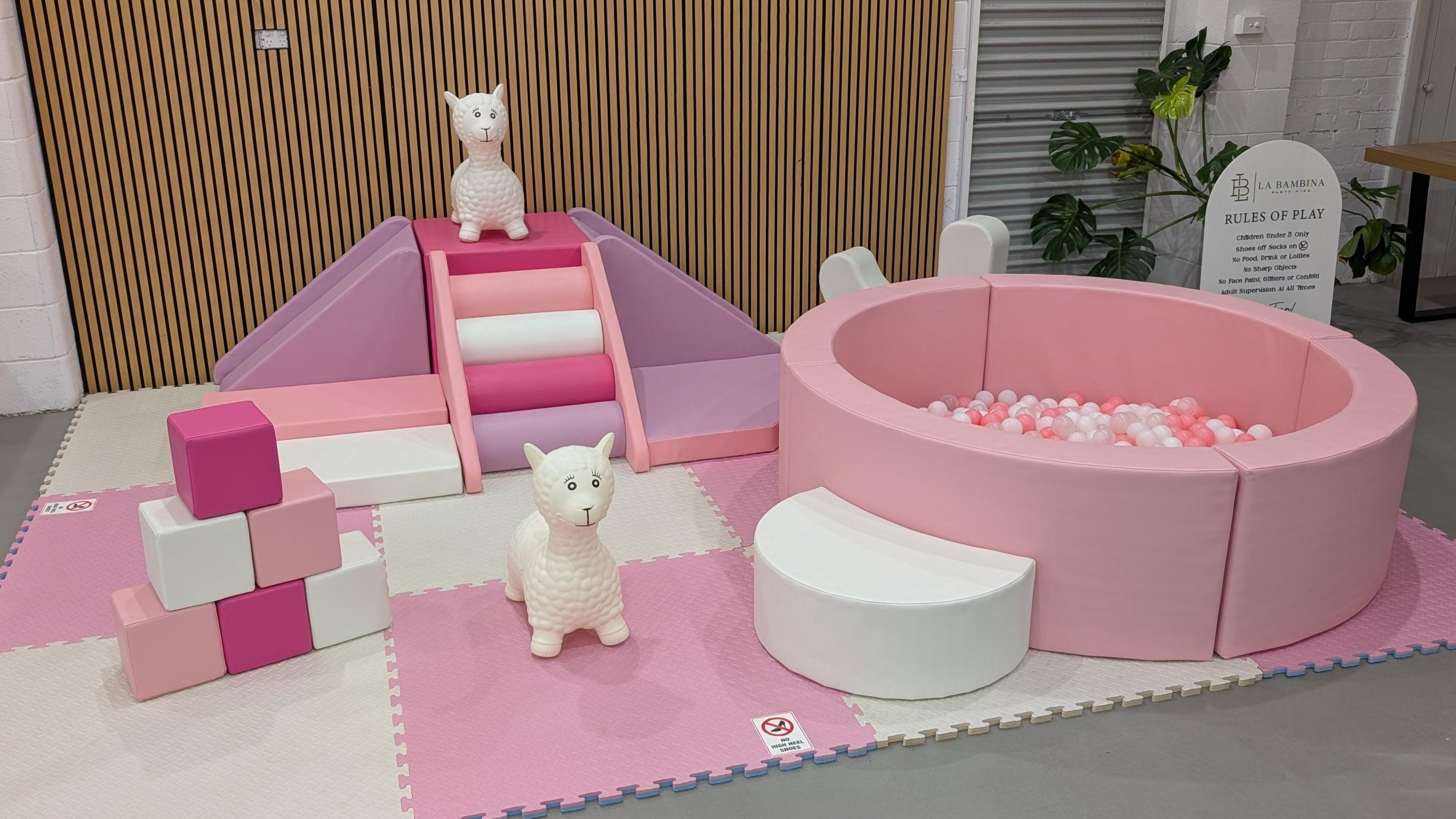 Pink and white soft play area with slide, ball pit, foam blocks, and llama toys on padded mats